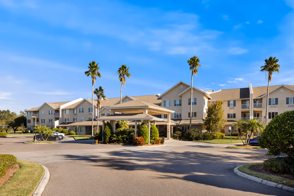 Exterior view of Azalea Park - A Provincial Senior Living Community, showing a large three-story building with beige walls and a tan roof. The entrance features a covered drop-off area surrounded by landscaping with palm trees and shrubs. The sky is clear and blue.