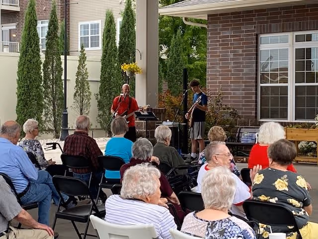A group of elderly people seated outdoors watching two musicians performing under a covered patio area. The setting includes tall green trees, a brick building, and a hanging flower basket.
