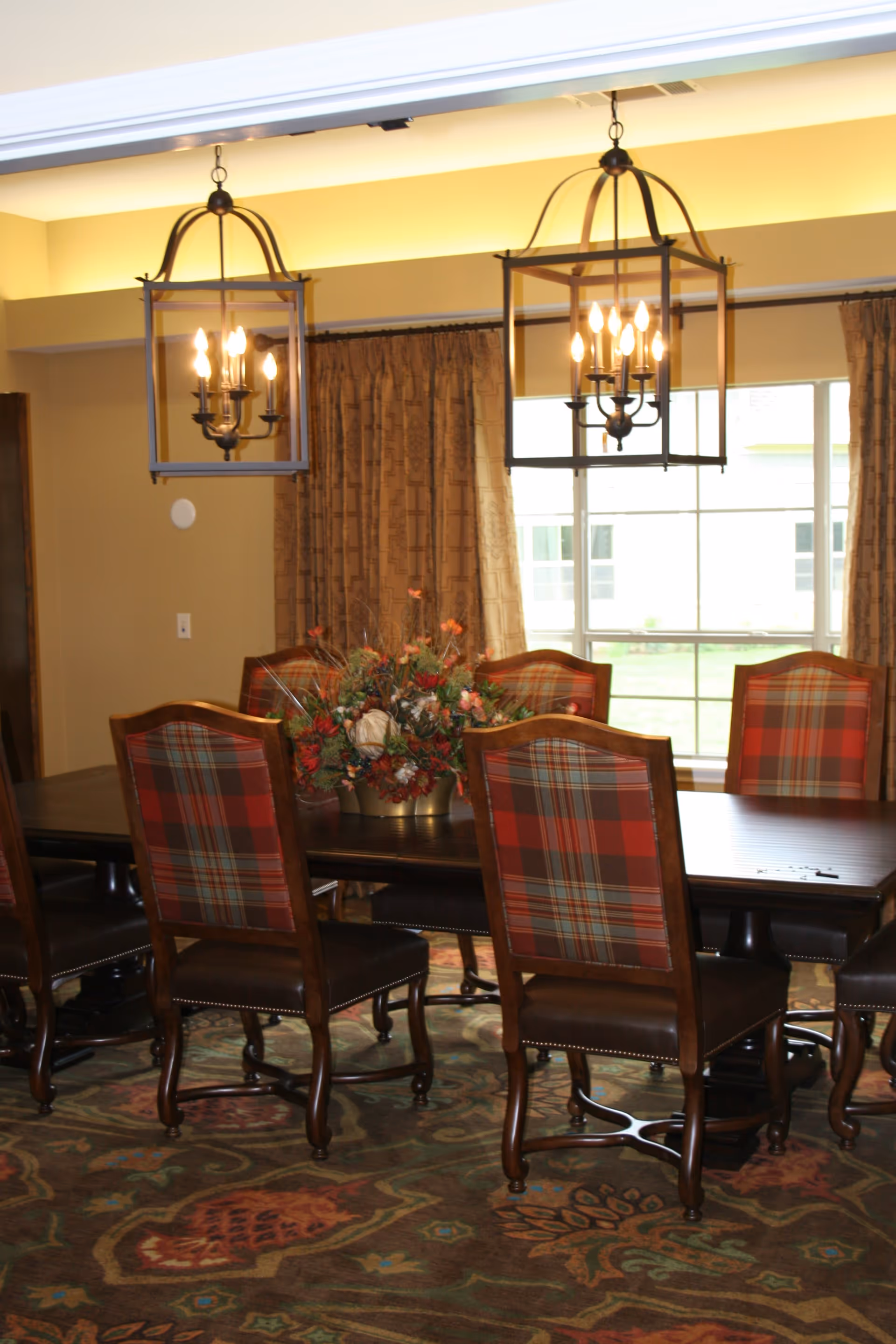 A dining room with a dark wooden table surrounded by chairs featuring red and green plaid upholstery. Two large, black metal lantern-style chandeliers hang from the ceiling above the table. A floral centerpiece decorates the middle of the table. The room has beige walls, a large window with brown curtains, and a patterned carpet.