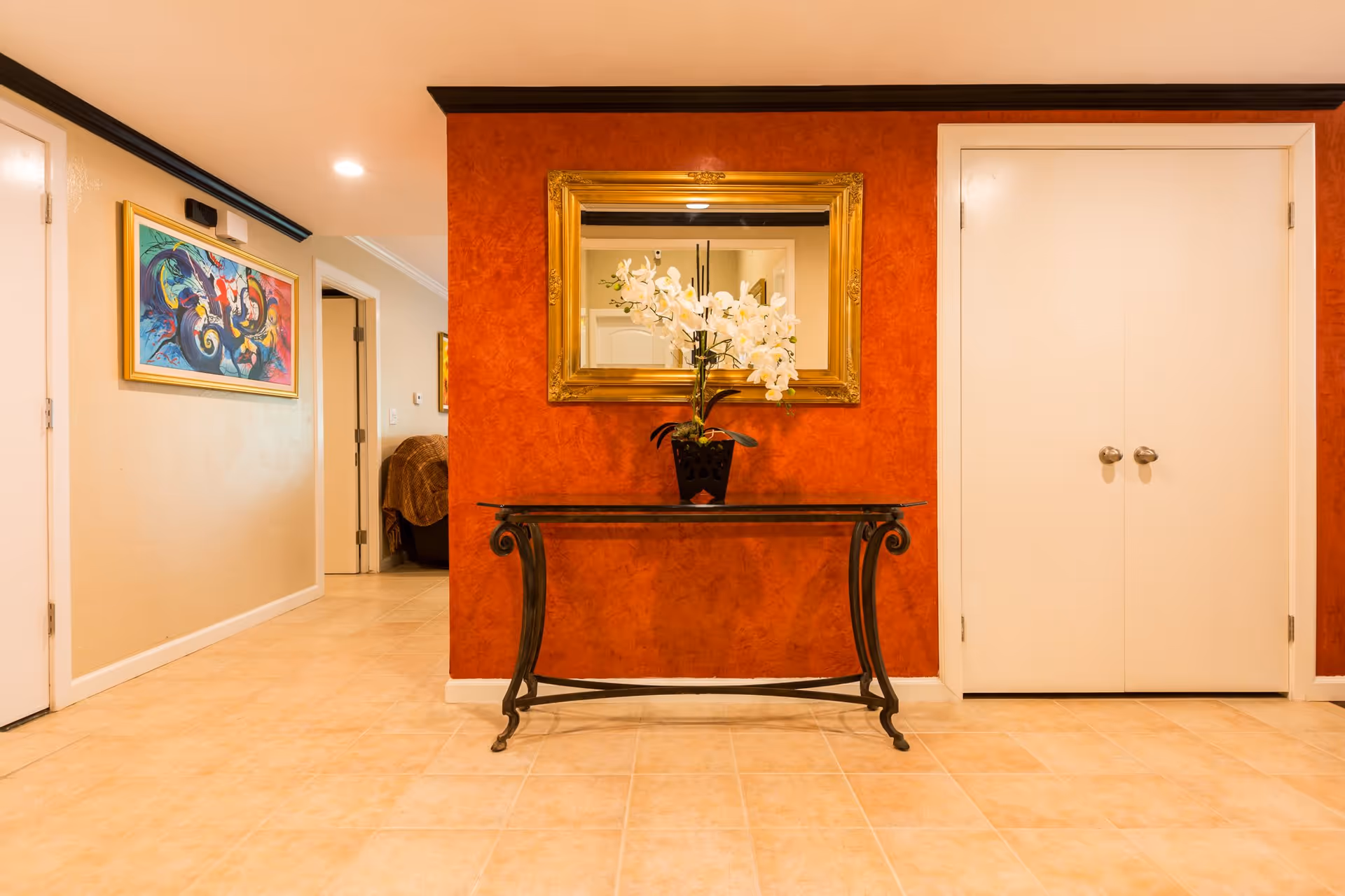 Interior hallway with beige tiled floor and walls painted in beige and burnt orange. A decorative black metal table with a glass top holds a black vase with white orchids. Above the table is a large gold-framed mirror. To the right is a set of white double doors, and to the left is a hallway with a colorful abstract painting on the wall and a glimpse of a room with a brown blanket on a chair.