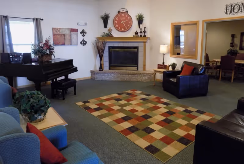Community lounge with a fireplace, piano, armchairs, and a multicolored checkered area rug.