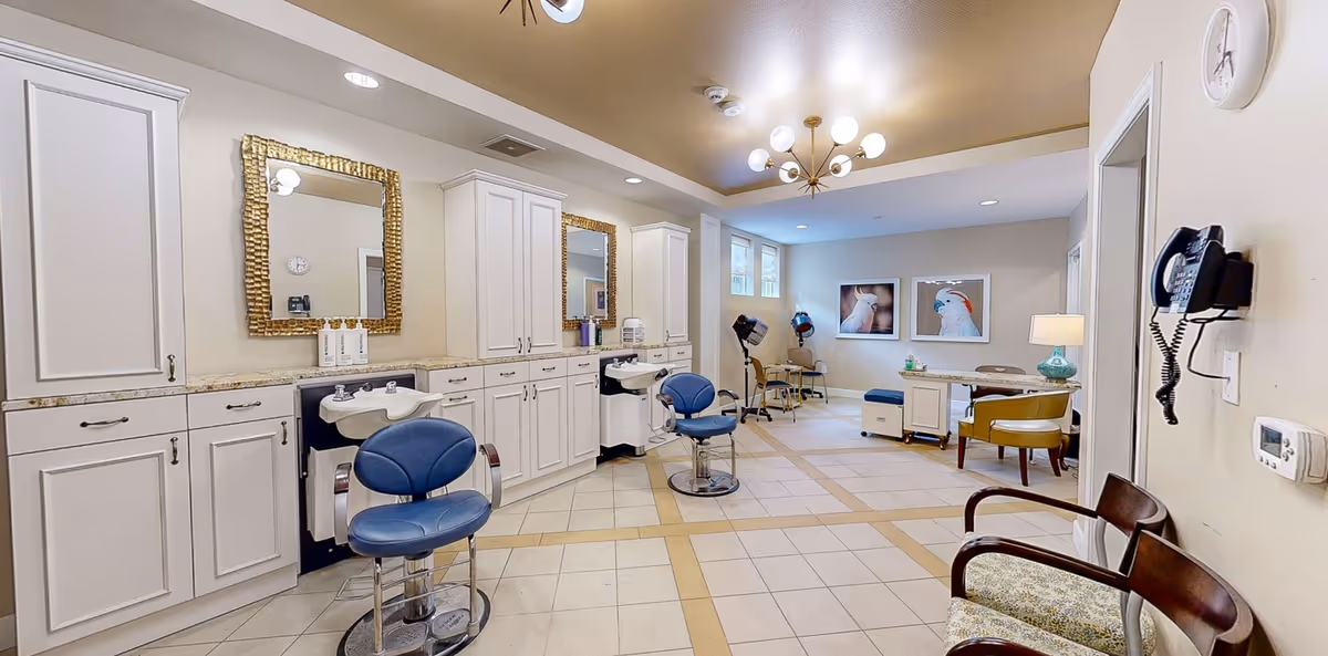 Interior view of a salon area in a senior living facility with white cabinetry, two blue salon chairs in front of sinks, hair drying chairs in the background, a desk with a lamp and chair, and two framed pictures of birds on the far wall.
