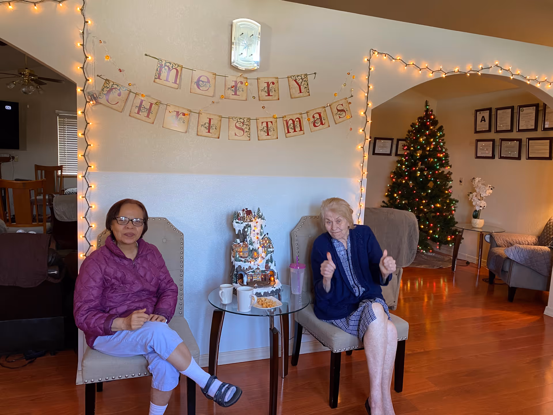 Two elderly women sitting on chairs in a warmly decorated living room with Christmas decorations including a lit Christmas tree, string lights, and a 'Merry Christmas' banner on the wall. A small glass table between them holds a decorative Christmas village, two mugs, and a plate of snacks.