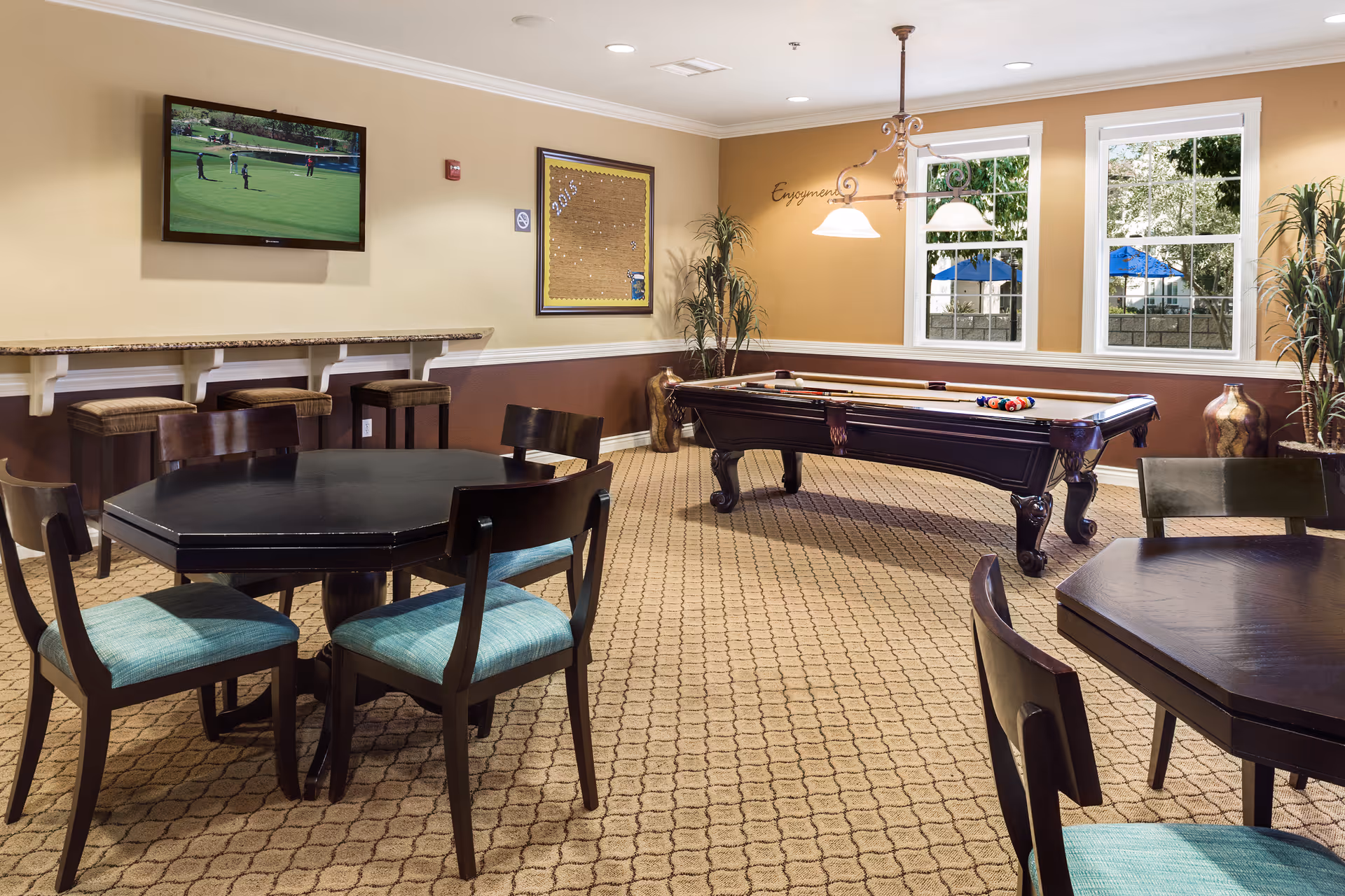 A recreational room with a pool table, several dark wood tables with teal cushioned chairs, a wall-mounted TV showing a golf game, and large windows letting in natural light. The room has beige walls with a darker brown lower half, carpeted floor, and decorative plants in the corners.