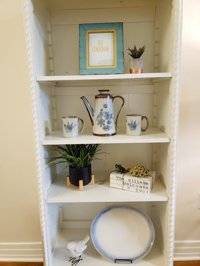 A white shelving unit with decorative items including a framed sign that says 'BE UNIQUE', a small potted plant, a ceramic coffee pot and two matching mugs with bird designs, a black pot with green grass-like plant, a small stack of books tied with string with a sign that says 'The Village welcomes You!', a decorative flower, a white ceramic bird figurine, and a large round plate with a blue rim.