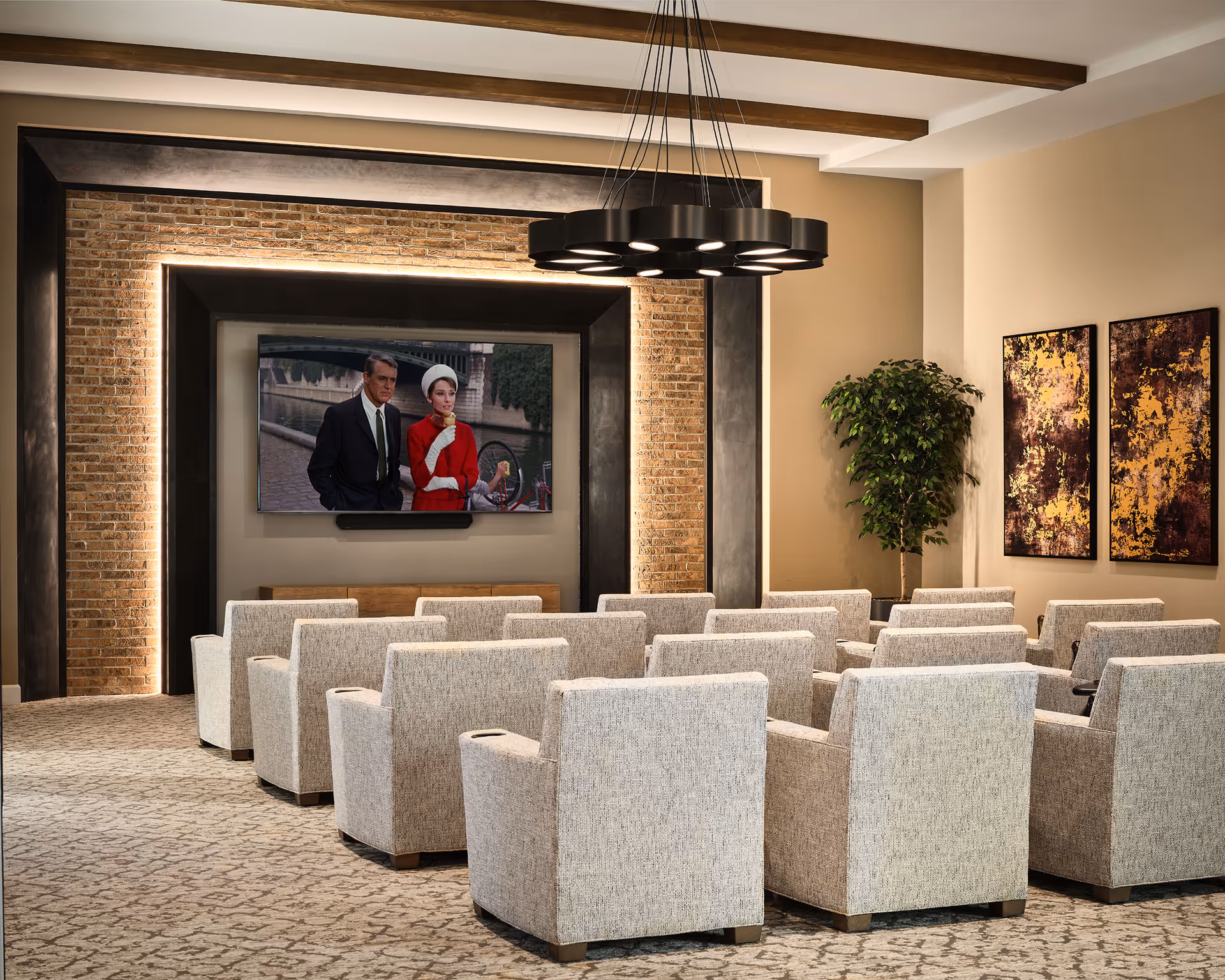 A cozy senior living facility media room with multiple rows of beige upholstered armchairs facing a large flat-screen TV mounted on a wall with decorative brick and black framing. The room features a modern circular ceiling light fixture, a potted plant in the corner, and two abstract paintings on the wall.