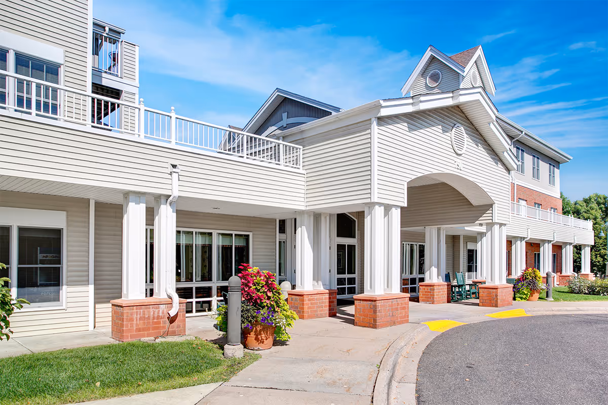 Exterior view of a senior living facility named Croixdale, featuring a covered entrance with white columns and brick bases, beige siding, multiple windows, and a clear blue sky above. There are potted plants with colorful flowers near the entrance and a curved driveway in front.