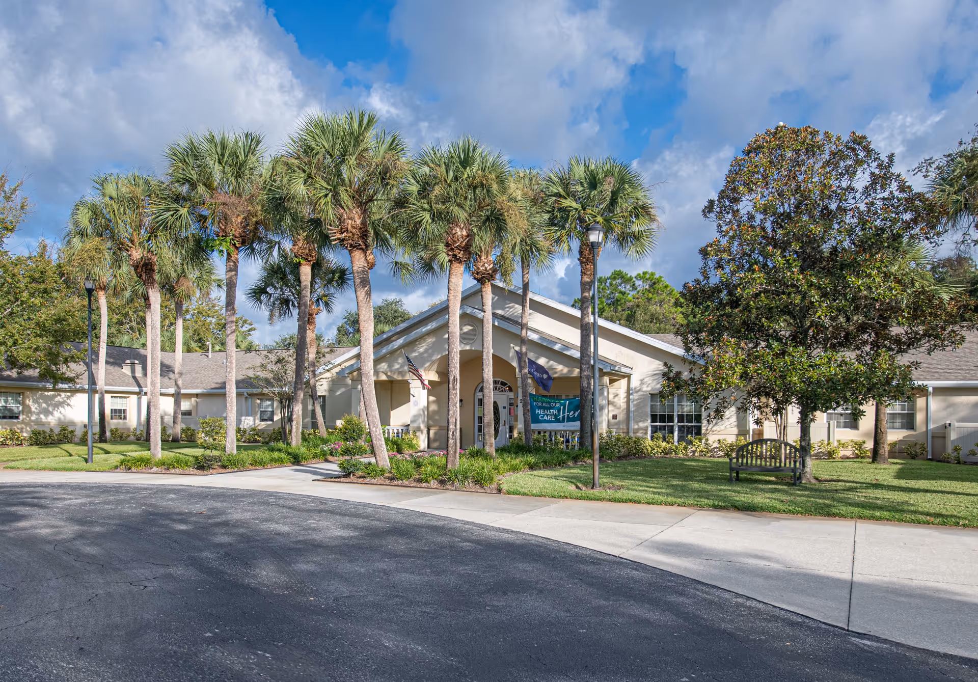 Exterior view of Arden Courts - ProMedica Memory Care Community in Palm Harbor, showing a single-story building with a covered entrance, surrounded by palm trees and well-maintained landscaping under a partly cloudy sky.