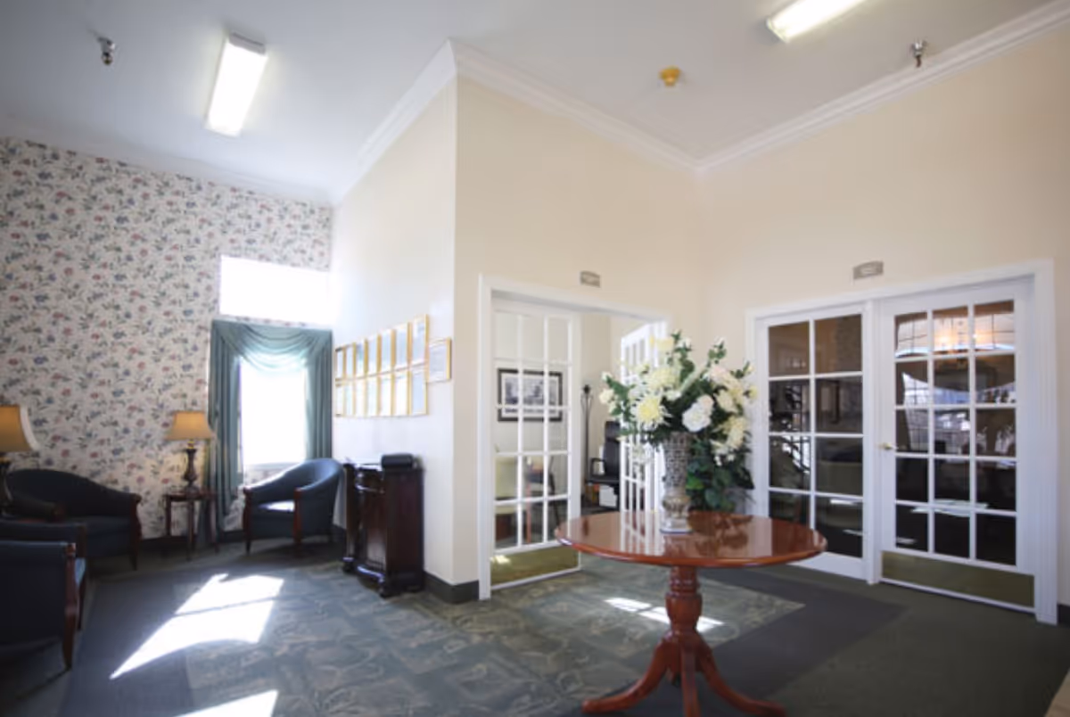 A senior living facility lobby with a round table topped by a large floral arrangement, seating area, and French doors.