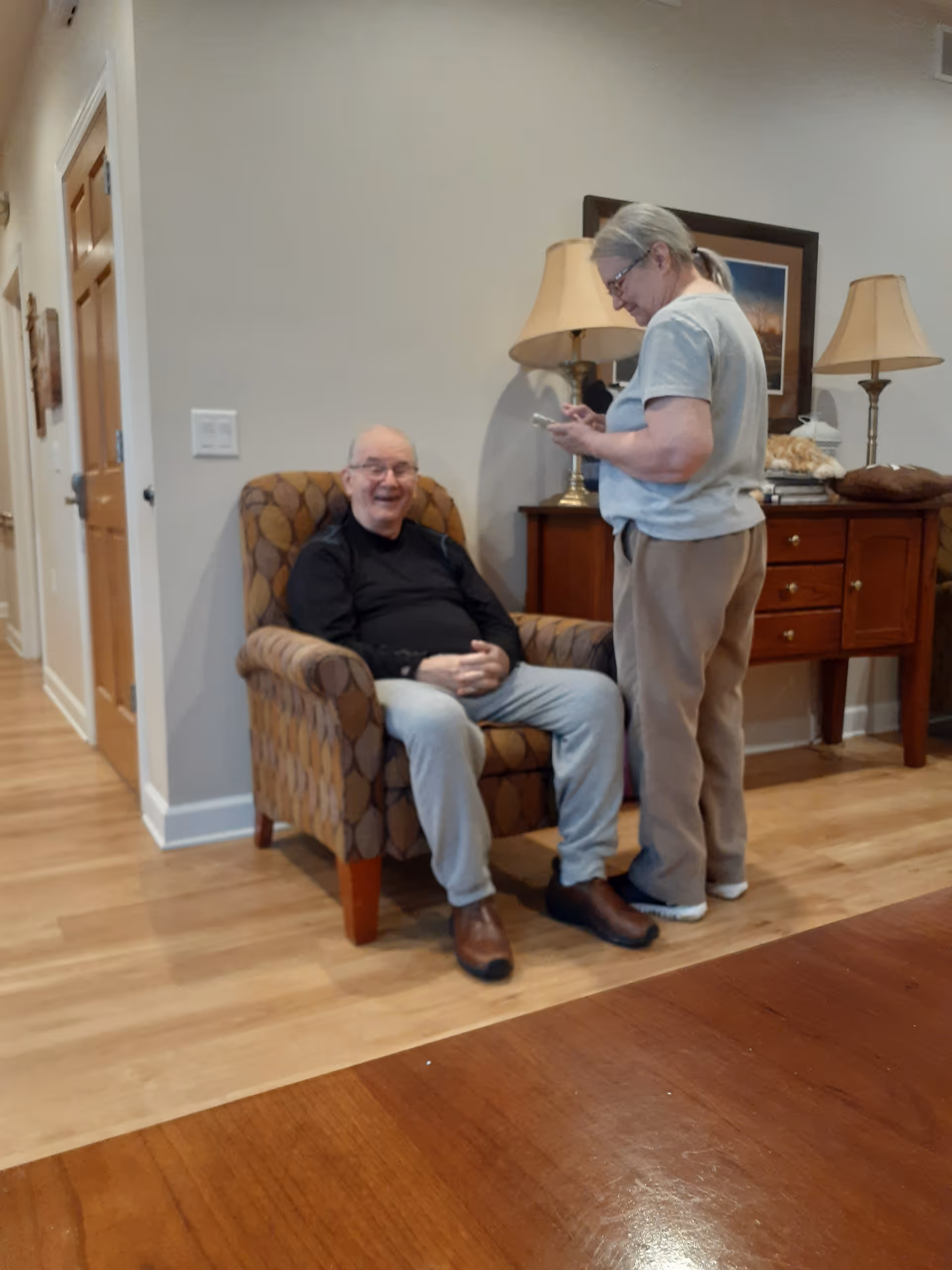 An elderly man sitting in a patterned armchair smiling, while a woman stands next to him looking at a smartphone. They are in a room with wooden floors, a wooden sideboard with two lamps, and a framed picture on the wall.