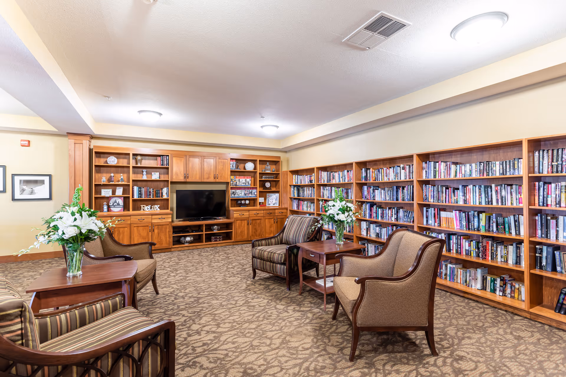 A cozy senior living community library with wooden bookshelves filled with books lining the walls. There are several comfortable armchairs arranged around small wooden tables with vases of white flowers. A large wooden entertainment center with a flat-screen TV and decorative items is visible at the far end of the room. The carpet has a patterned design, and the ceiling has recessed lighting.