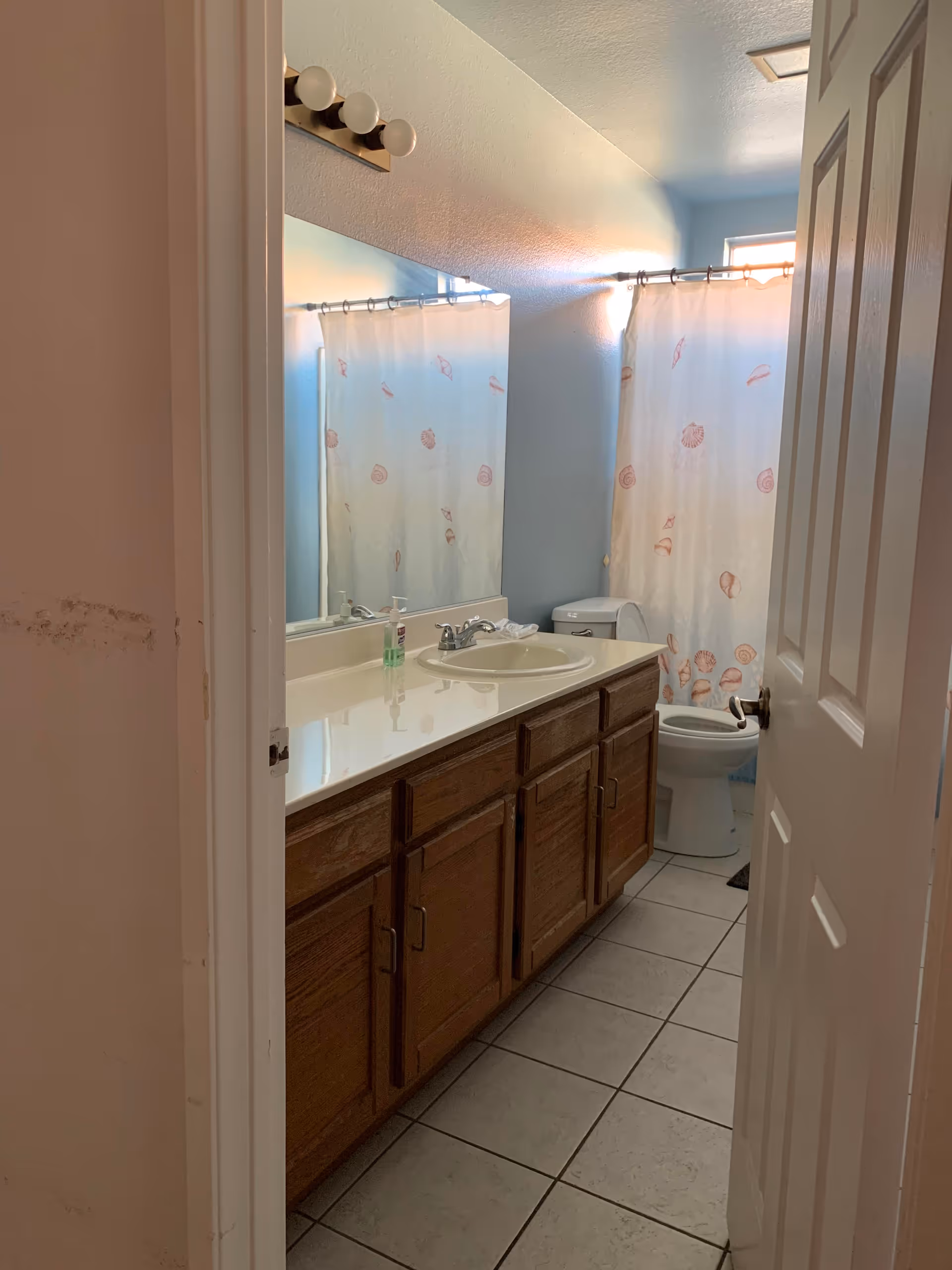 View into a bathroom with a long vanity and sink over wooden cabinets, tiled floor, toilet, and a shower with a seashell-patterned curtain.
