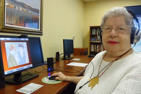 An elderly woman wearing glasses and headphones is seated at a desk using a computer mouse. On the computer screen, there is an illustration of two people and some text options. The room has light yellow walls, a framed picture, bookshelves, and another computer monitor in the background.
