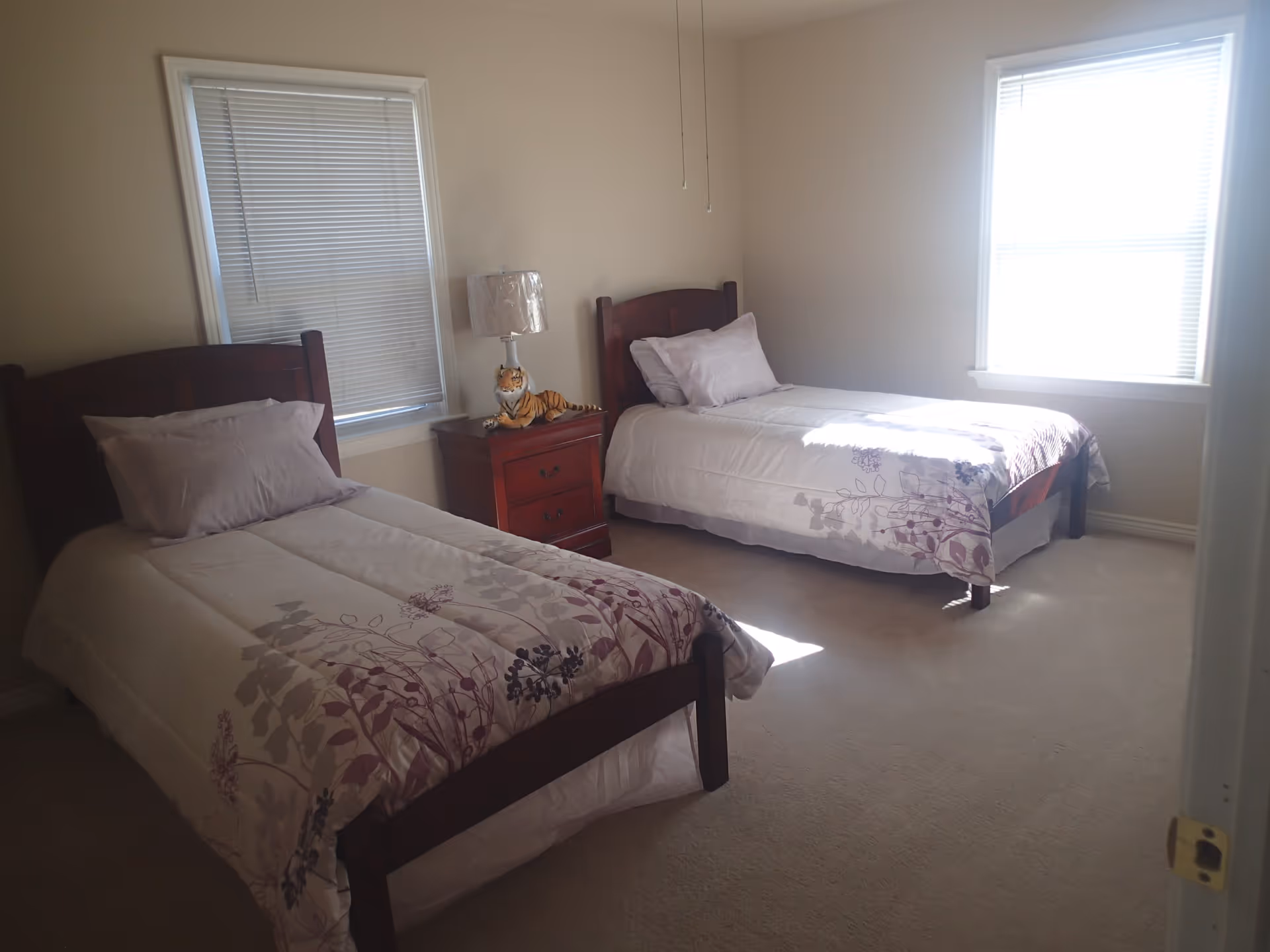 A bright bedroom with two twin beds, a nightstand and lamp between them, and windows with blinds.