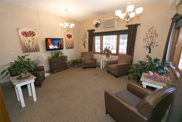 A cozy living room with four armchairs arranged around the perimeter, two small white side tables with potted plants, a flat-screen TV mounted on the wall, and decorative wall art featuring flowers and trees. The room has beige walls, a carpeted floor, a window with brown curtains, and ceiling light fixtures.