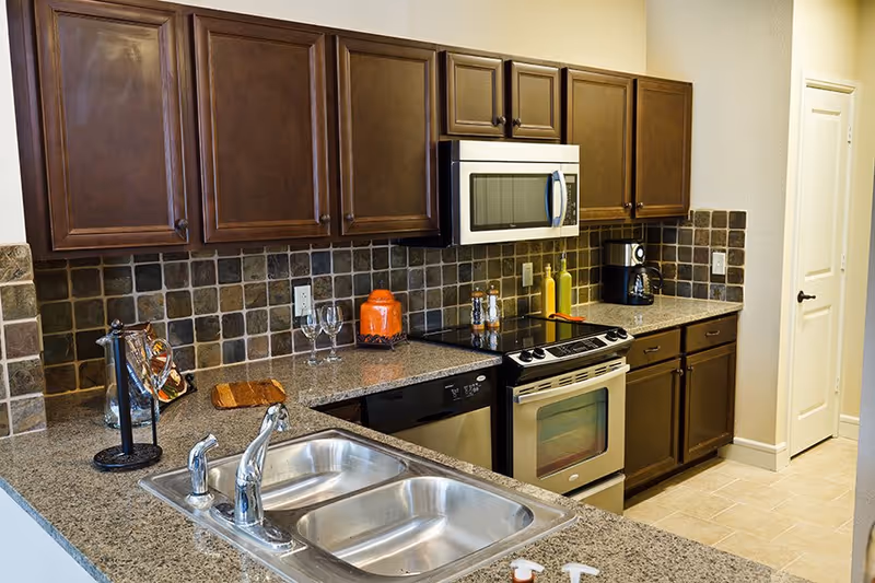 Modern kitchen with granite countertops, a double stainless steel sink, dark wooden cabinets, a stainless steel oven with a stovetop, a microwave above the oven, a dishwasher, a coffee maker, and various kitchen items including two wine glasses, a cutting board, and condiment bottles.
