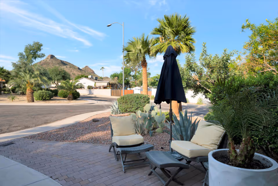 Outdoor patio area with cushioned chairs, a small table, and a closed black umbrella surrounded by desert plants and palm trees, with a mountain visible in the background under a clear blue sky.