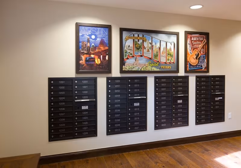 Interior view of a senior living facility mailroom with four sets of black mailboxes mounted on a beige wall. Above the mailboxes are three framed posters featuring Austin, Texas themes, including cityscapes and a guitar shop advertisement. The floor is wooden and there is a small wooden table partially visible on the left.