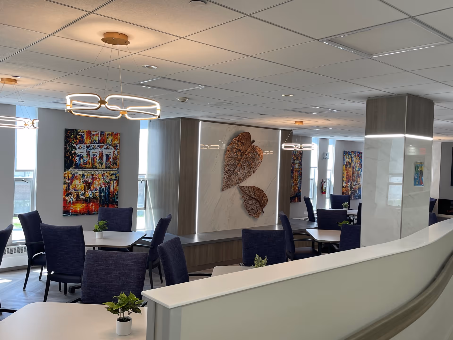 A modern dining area with multiple tables and dark blue chairs. The room features contemporary light fixtures hanging from the ceiling, colorful abstract paintings on the walls, and decorative metal leaf art on a marble panel. Small potted plants are placed on the tables, and large windows allow natural light to brighten the space.