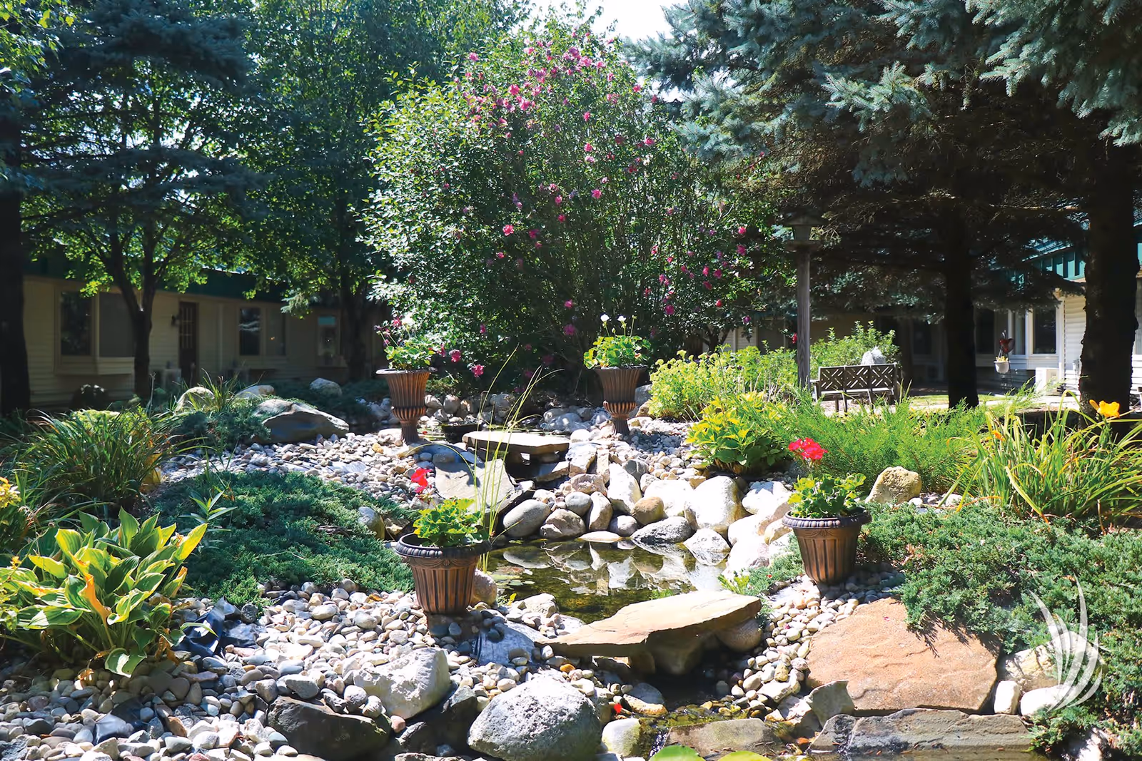 A peaceful outdoor garden area at Valley Pines Senior Living featuring a small pond with rocks and potted plants around it, surrounded by lush greenery and trees with a building visible in the background.