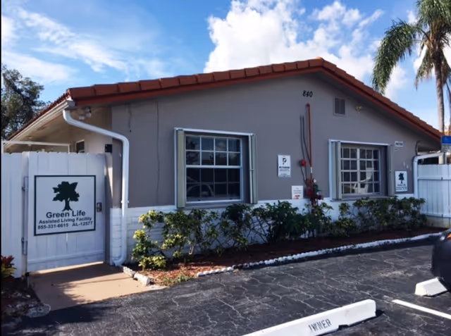 Exterior view of a single-story building with a tiled roof and two windows. There is a white gate on the left side with a sign that reads 'Green Life Assisted Living Facility' along with a phone number. The building is surrounded by small bushes and a parking area with marked parking spaces in front. A palm tree is visible on the right side under a partly cloudy sky.