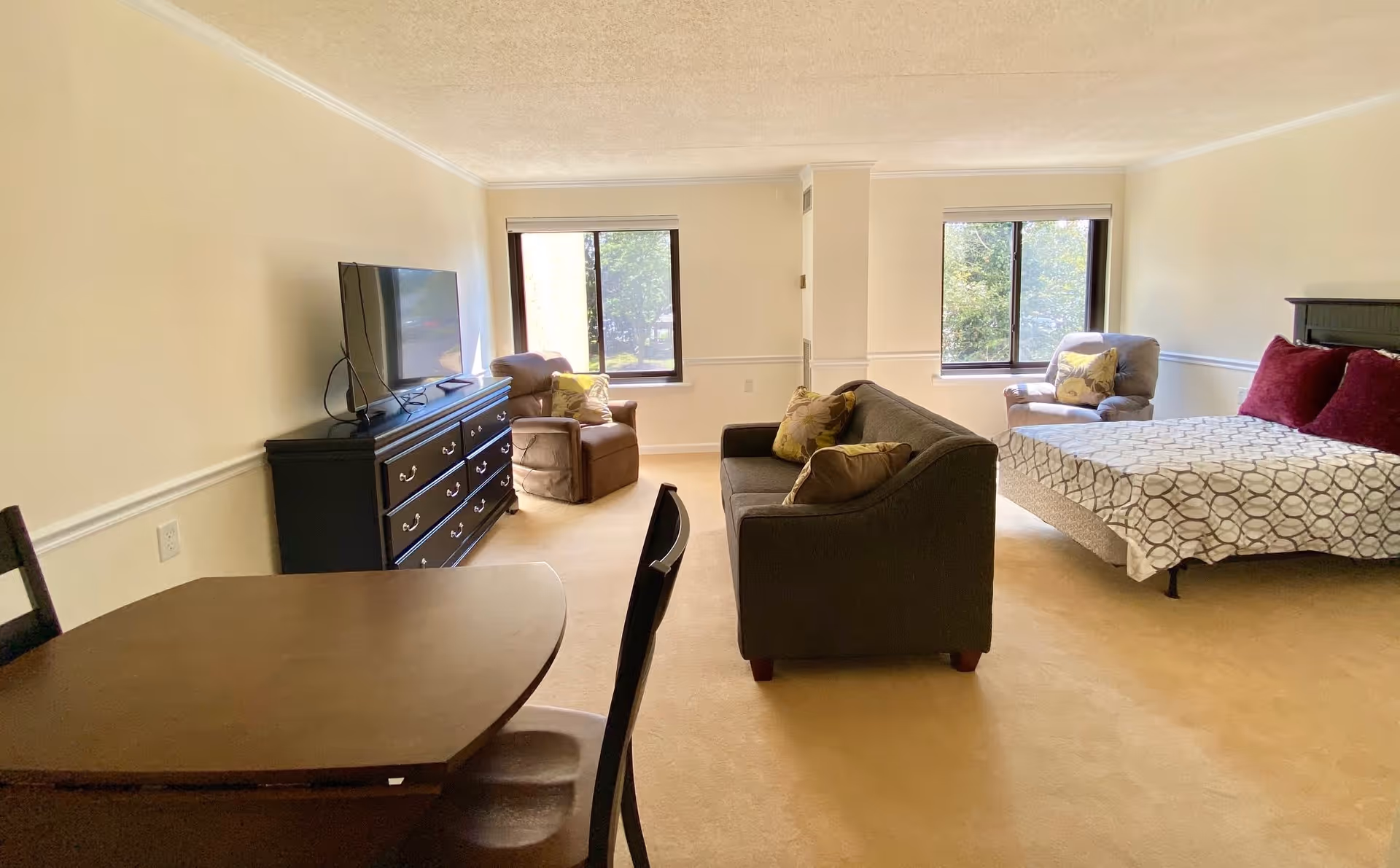 A spacious, well-lit room with beige walls and carpet featuring a bed with patterned bedding and red pillows, a dark gray sofa with yellow pillows, two armchairs near windows, a dark wooden dresser with a flat-screen TV on top, and a wooden dining table with chairs in the foreground.