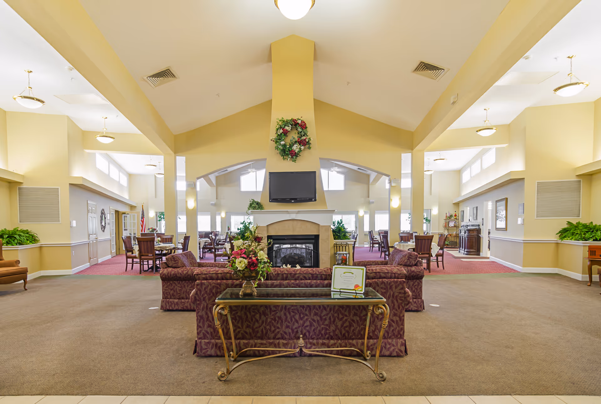 Spacious and well-lit common area in an assisted living facility featuring a central fireplace with a TV mounted above it, floral wreath decoration, and comfortable seating including patterned sofas and a glass-top table with a flower arrangement. Surrounding the fireplace are multiple dining tables and chairs, with large windows allowing natural light to fill the room. The walls are painted in soft yellow tones and the ceiling is vaulted with several light fixtures.