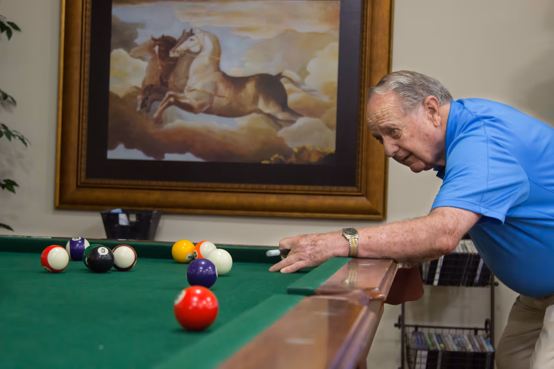 An elderly man in a blue shirt is playing pool indoors, aiming to strike the cue ball on a green pool table. Behind him, there is a framed painting of horses running through clouds on the wall.