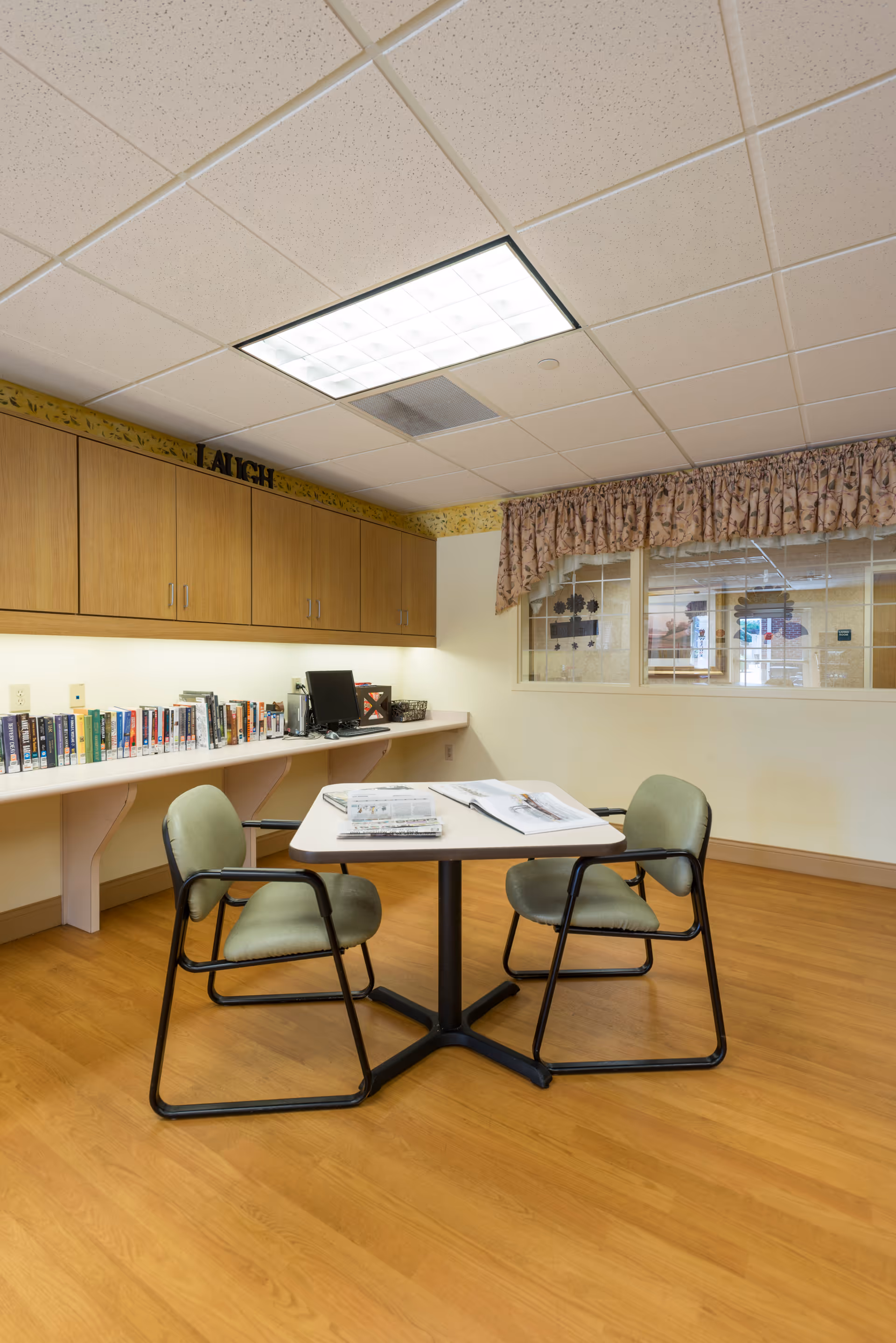A small room with a square table and two green cushioned chairs with black metal frames. On the table are newspapers and magazines. Along one wall is a long white counter with a row of books, a computer, and wooden cabinets above. The room has wood flooring, a drop ceiling with fluorescent lighting, and a window with floral curtains looking into another room.