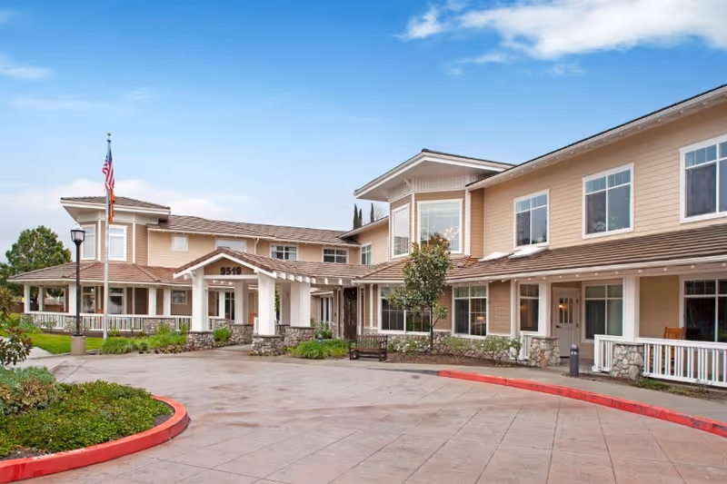 Exterior view of a two-story senior living facility building with beige siding and stone accents. The entrance features a covered porch with white pillars and a sign displaying the number 9519. There is a flagpole with an American flag near the entrance, surrounded by landscaped greenery and a paved driveway with a red curb.