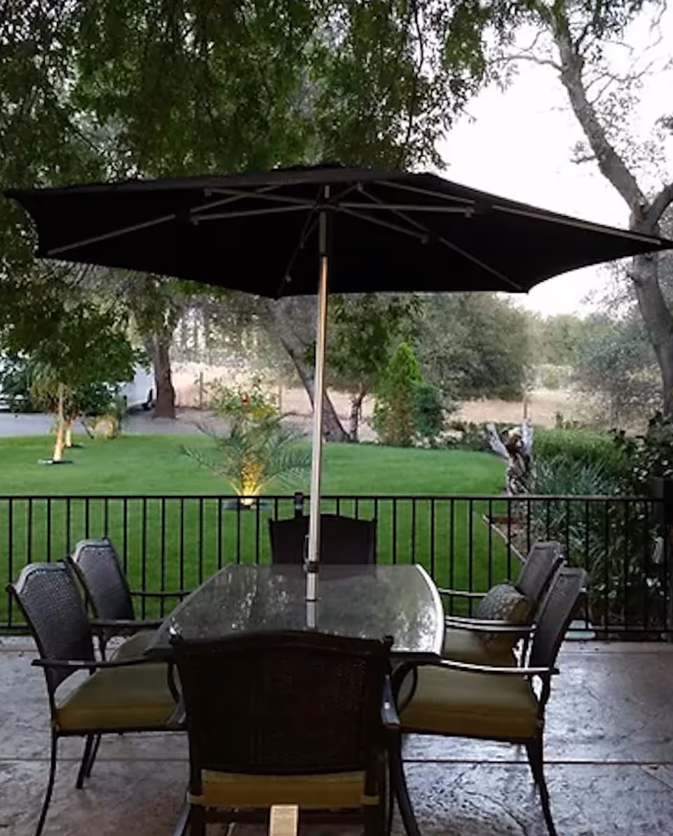 Outdoor patio area with a glass-top table surrounded by six cushioned chairs and a large black umbrella overhead. The patio overlooks a green lawn with trees and shrubs in the background.