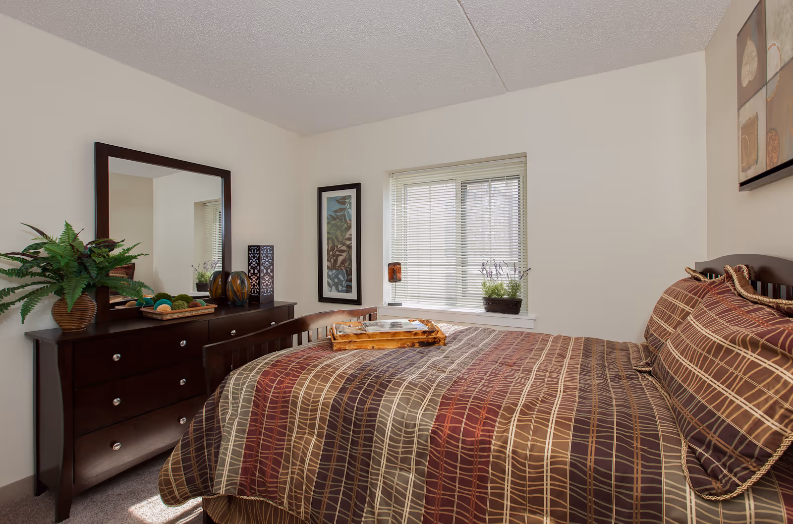 A cozy bedroom with a bed covered in a brown, red, and beige plaid patterned comforter and matching pillows. A wooden dresser with a large mirror stands against the wall, decorated with a potted plant, decorative balls, and vases. A window with blinds lets in natural light, and a framed artwork hangs on the wall next to the window. A small tray is placed on the bed.