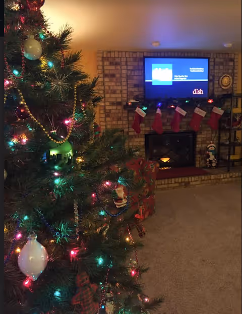 Decorated Christmas tree in a living room beside a brick fireplace with stockings hung and a TV above.