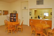 Communal senior living room with round wooden tables and chairs, a service window with counter, bookshelf and bulletin board on the wall.