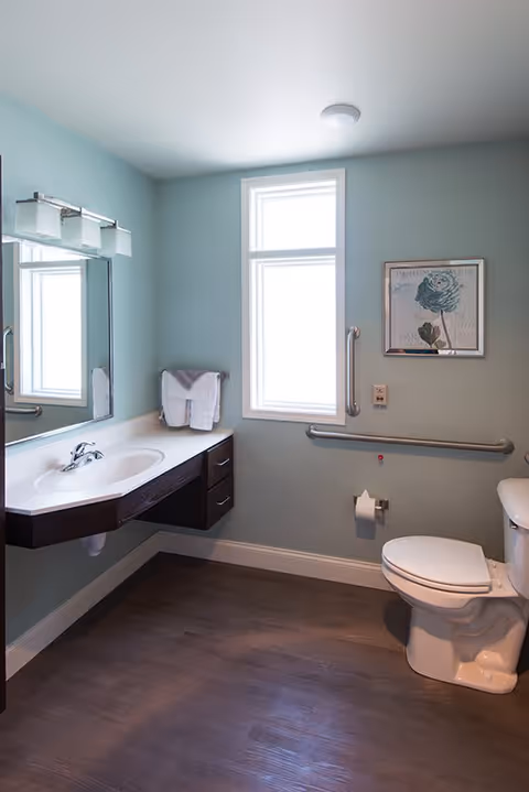 A clean and accessible bathroom with light blue walls, a white toilet, a wall-mounted sink with a dark wood cabinet, a large mirror above the sink, a window letting in natural light, grab bars on the walls, a towel rack with folded towels, and a framed floral artwork.
