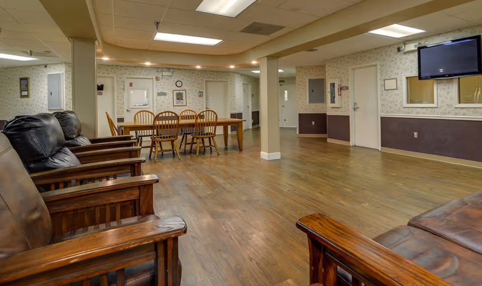 Interior view of a senior living facility common area with wooden armchairs and a leather sofa on the left, a wooden dining table with chairs in the background, wood flooring, wallpapered walls, and a wall-mounted TV on the right.