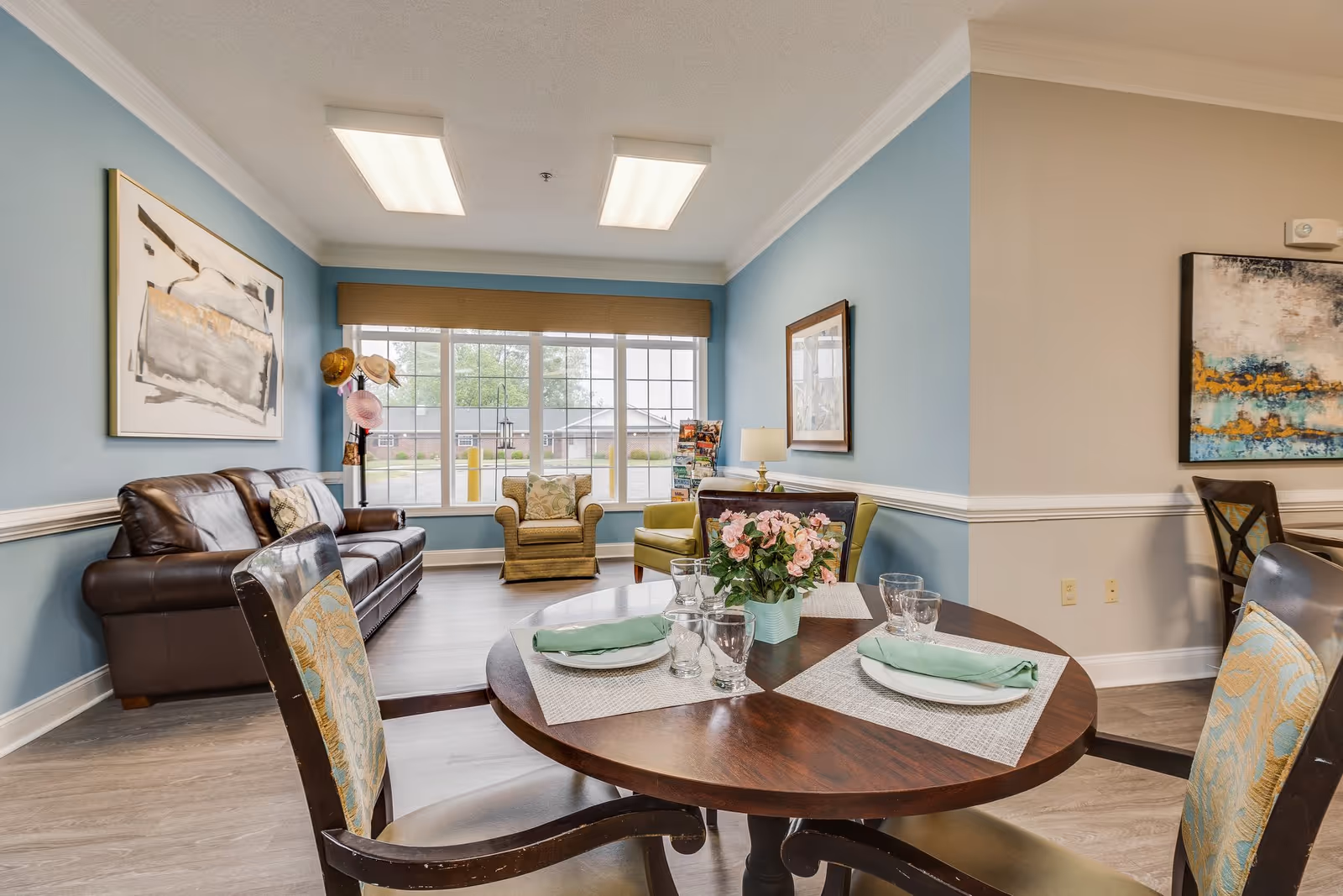 A cozy living and dining area in an assisted living facility with a round wooden table set for two, featuring plates, glasses, and green napkins. The room has light blue walls, a large window with a view outside, a brown leather sofa, two armchairs, and framed artwork on the walls. A vase with pink flowers is placed on the dining table.