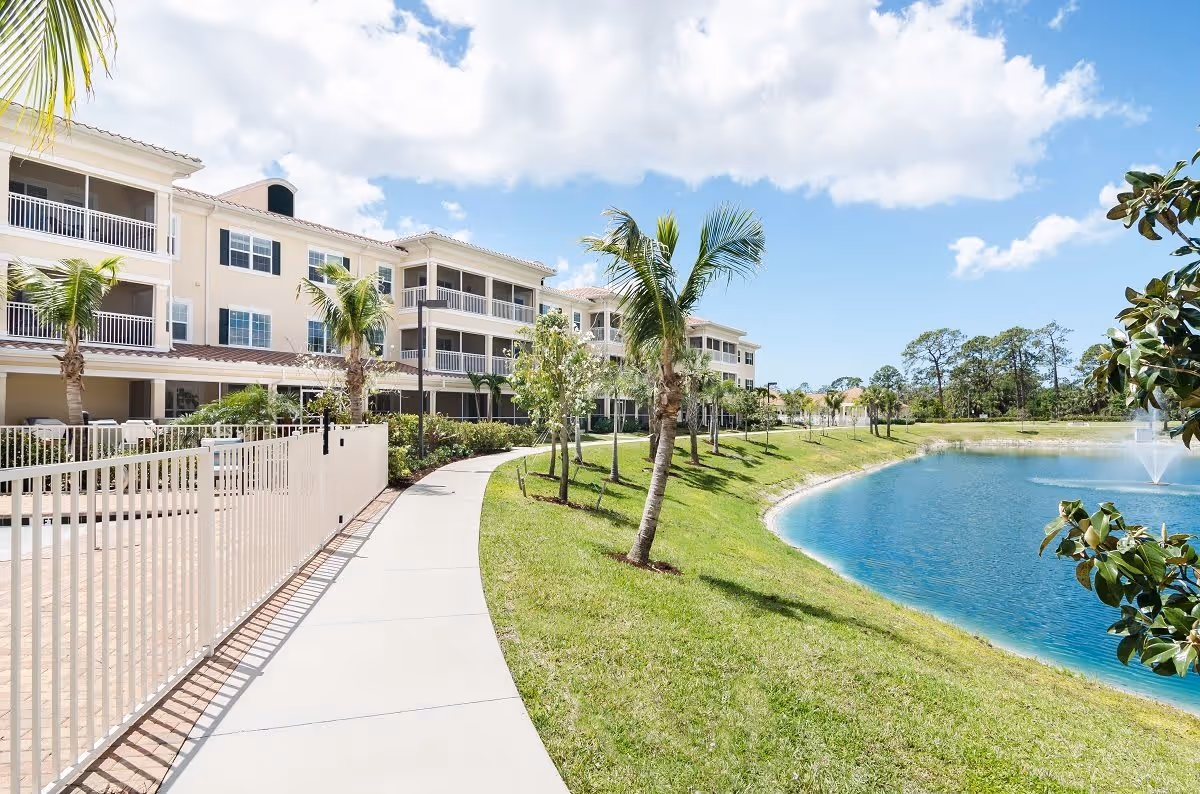 A sunny outdoor scene at American House Bonita Springs featuring a paved walkway lined with palm trees and a white fence on the left side. To the right, there is a grassy area sloping down to a pond with a water fountain. In the background, there is a three-story residential building with balconies and windows under a partly cloudy blue sky.