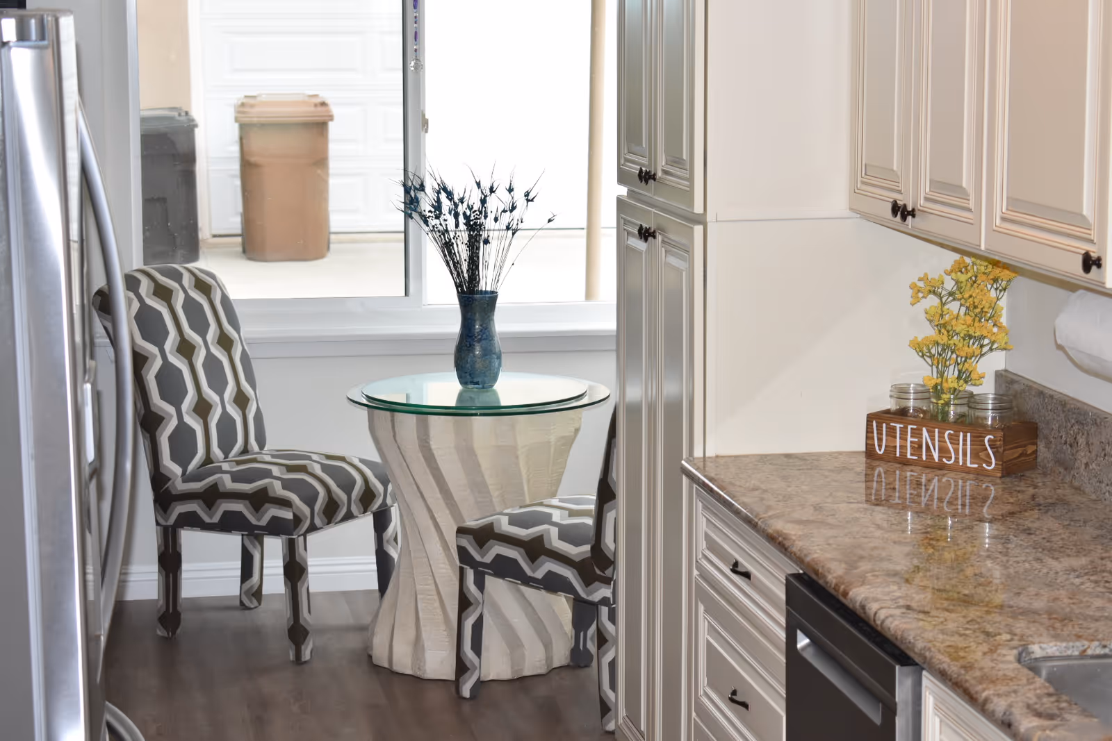 A small kitchen nook with two patterned chairs and a round glass-top table with a blue vase holding decorative branches. The kitchen has cream-colored cabinets, a granite countertop with a wooden box labeled 'UTENSILS' holding yellow flowers, and stainless steel appliances. A window behind the table shows two trash bins outside.