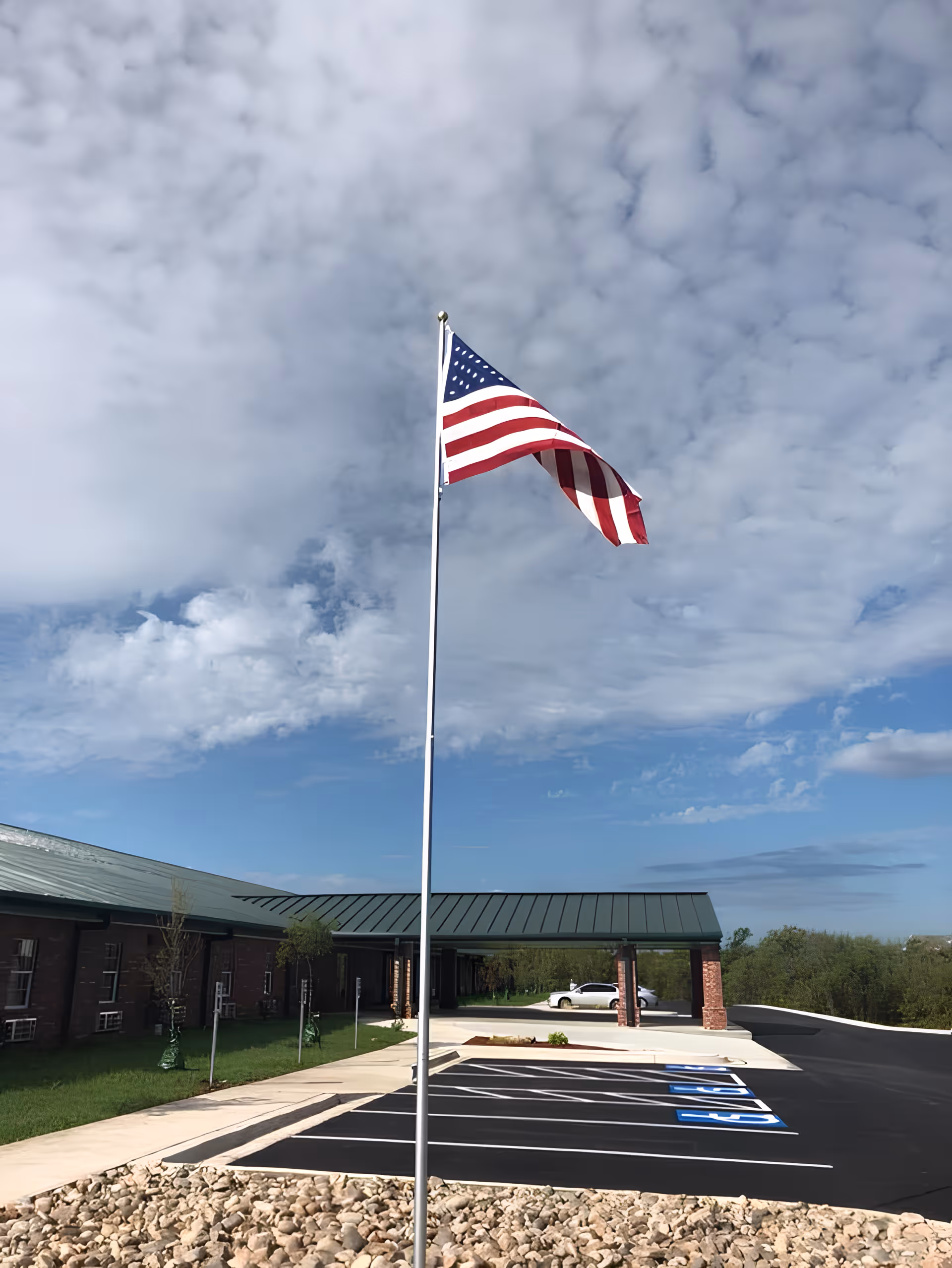 An American flag on a tall pole in front of a single-story brick building and parking lot under a cloudy sky.