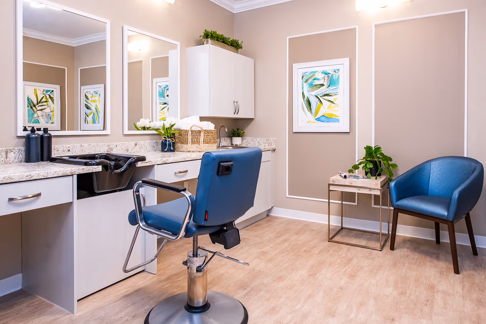 A bright and clean salon area with a blue salon chair in front of a countertop with a sink, two large mirrors, and cabinets. There is a small table with a plant and a blue armchair against the beige wall decorated with framed artwork and white trim.