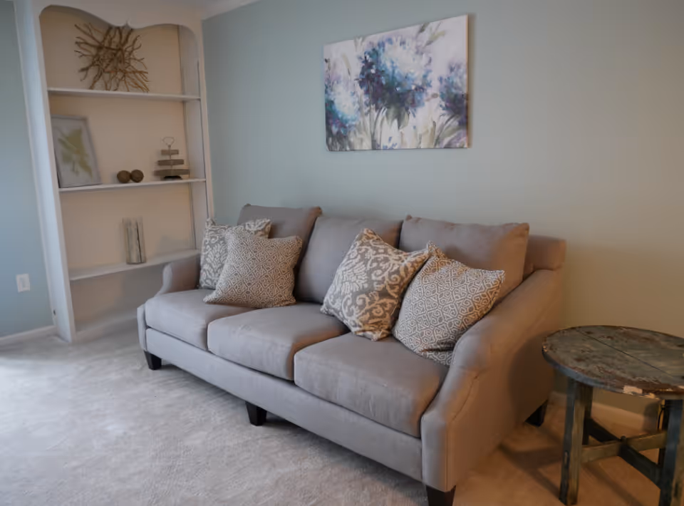 Gray upholstered sofa with patterned throw pillows in a light living room with built-in shelving, a floral painting, and a small round side table.
