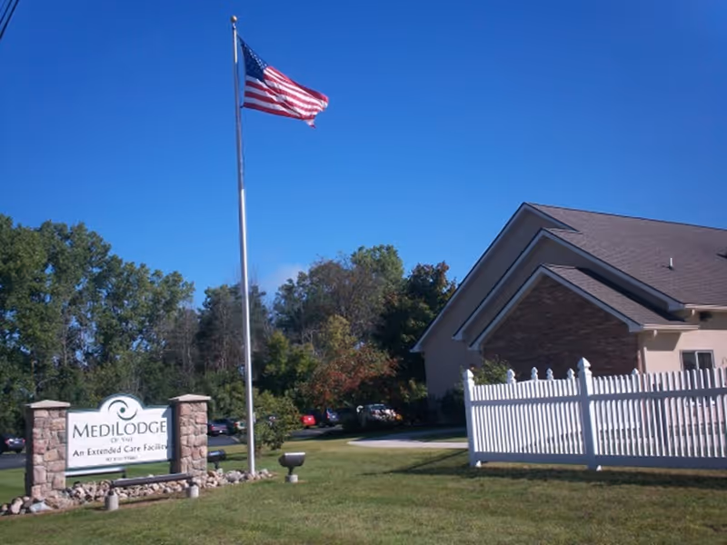 Exterior view of Medilodge of Yale, an extended care facility, showing a stone sign with the facility name, an American flag on a tall flagpole, a white picket fence, part of the building, and trees in the background under a clear blue sky.