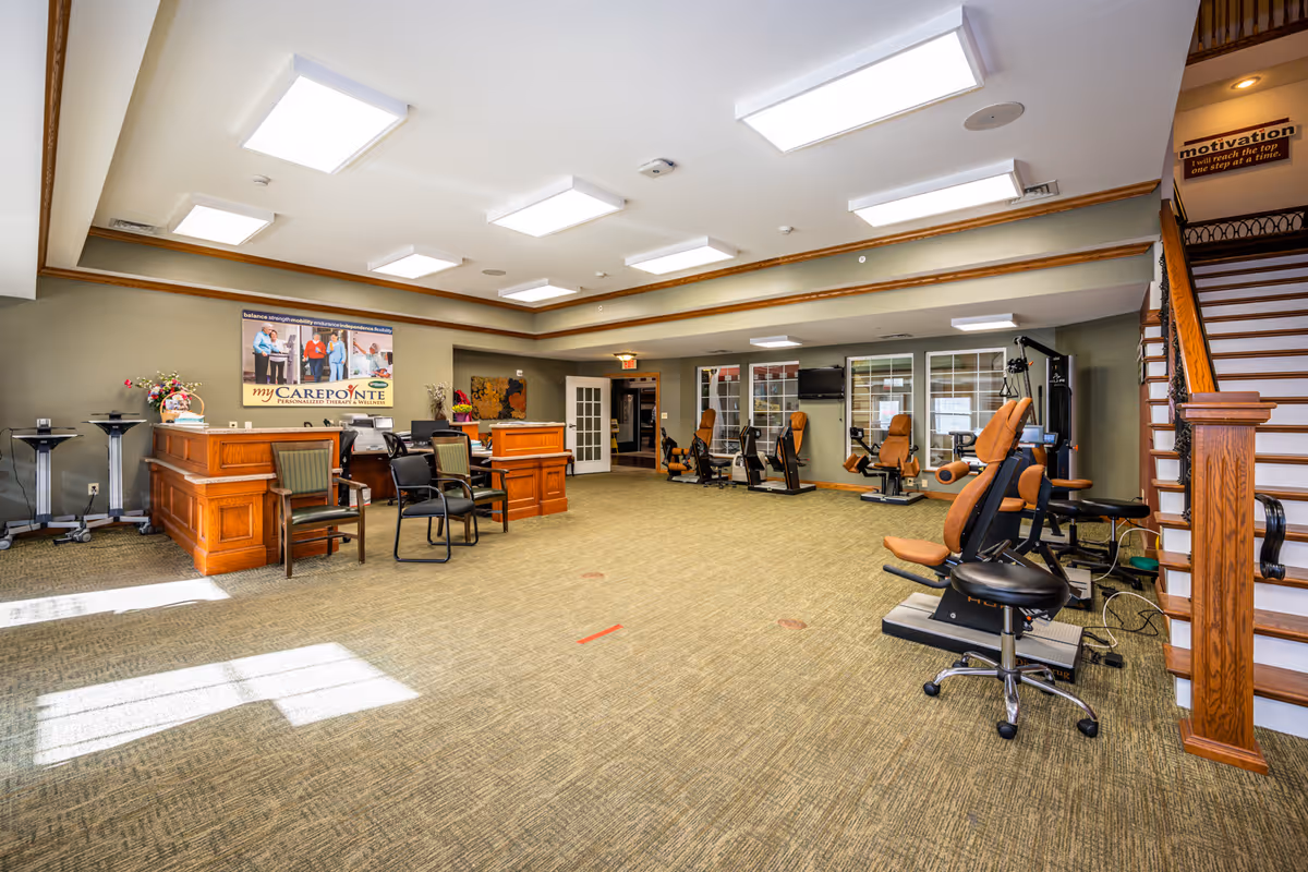 A spacious rehabilitation and wellness room with exercise equipment including stationary bikes and weight machines. There is a wooden reception desk with chairs in front, a staircase with wooden railing on the right, and motivational posters on the walls. The room is well-lit with ceiling lights and natural light from windows.