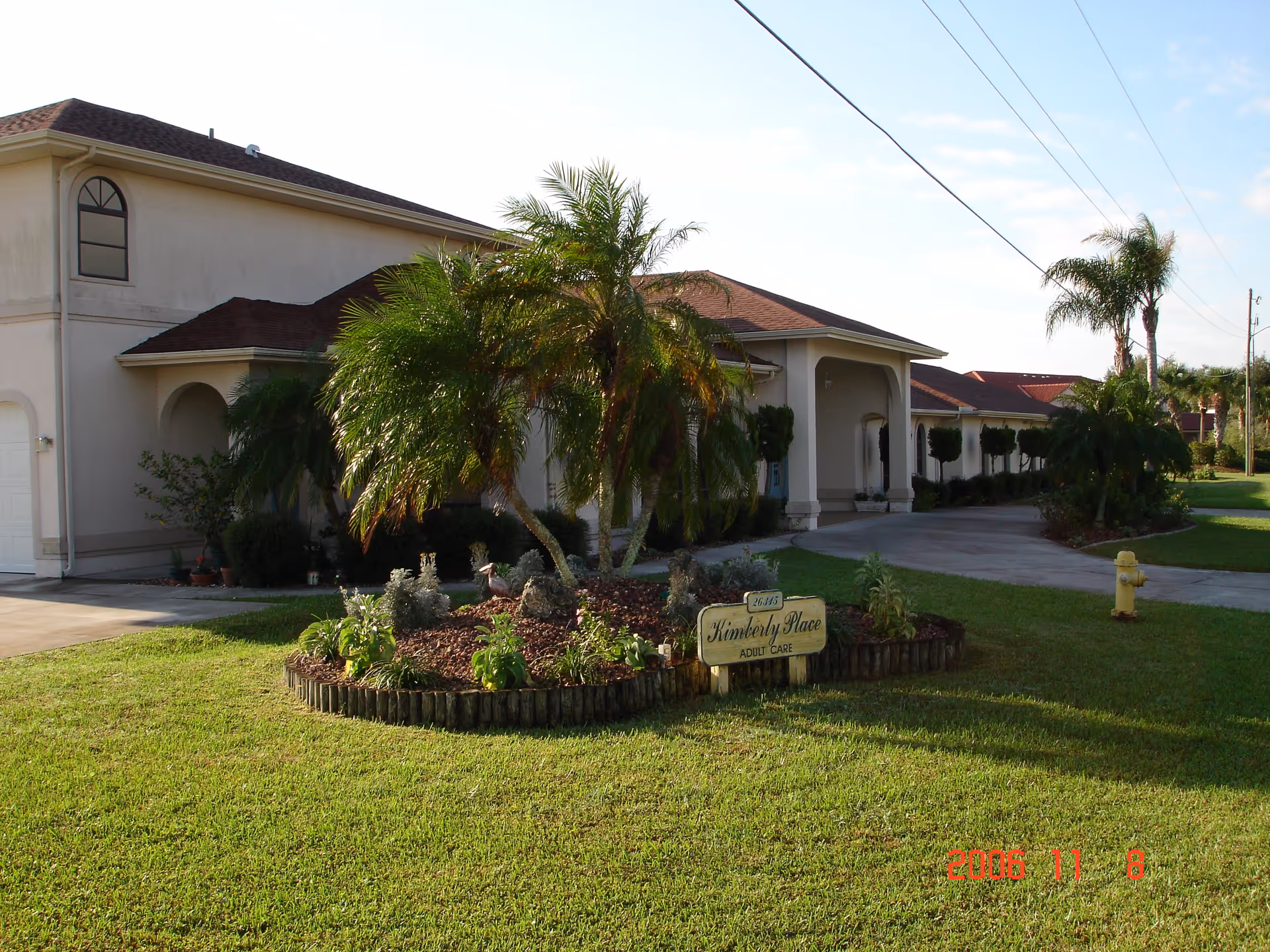 Exterior view of Kimberly Place adult care facility showing a beige building with a red roof, a landscaped garden with palm trees and plants, a driveway, and a yellow fire hydrant on a well-maintained lawn under a clear sky.