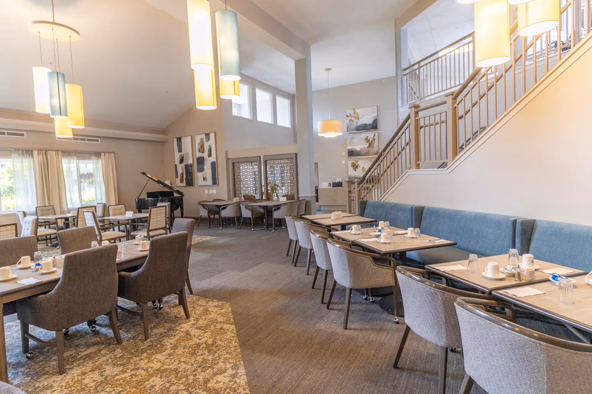 A spacious dining room with multiple tables and chairs arranged neatly. The tables are set with cups, glasses, and napkins. There is a blue cushioned bench along one wall and several modern pendant lights hanging from the ceiling. A staircase with wooden railings is visible on the right side, and large windows allow natural light to fill the room. Artwork is displayed on the walls, and a piano is seen in the background near the windows.