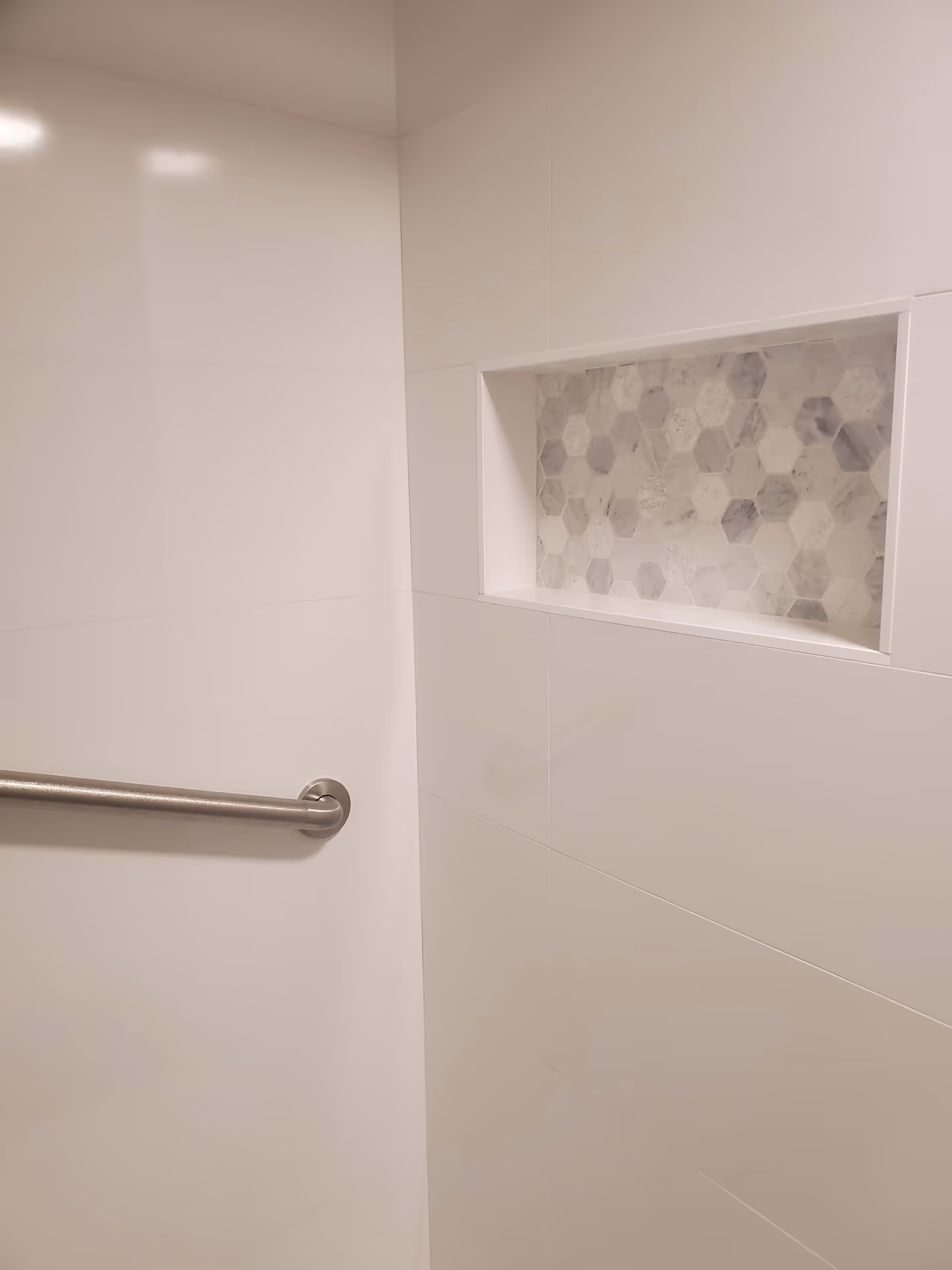 A close-up view of a shower wall with white tiles and a built-in niche featuring hexagonal patterned tiles in shades of gray and white. A stainless steel grab bar is mounted on the left side of the wall.