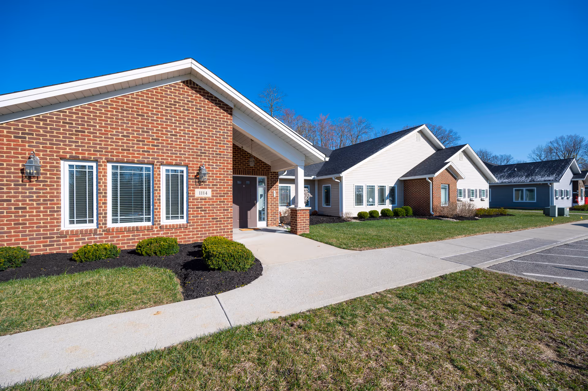 Exterior view of a senior living facility building with a combination of red brick and white siding. The building has multiple windows with white frames, a covered entrance with a brown door, and neatly trimmed bushes along the walkway. The sky is clear and blue.