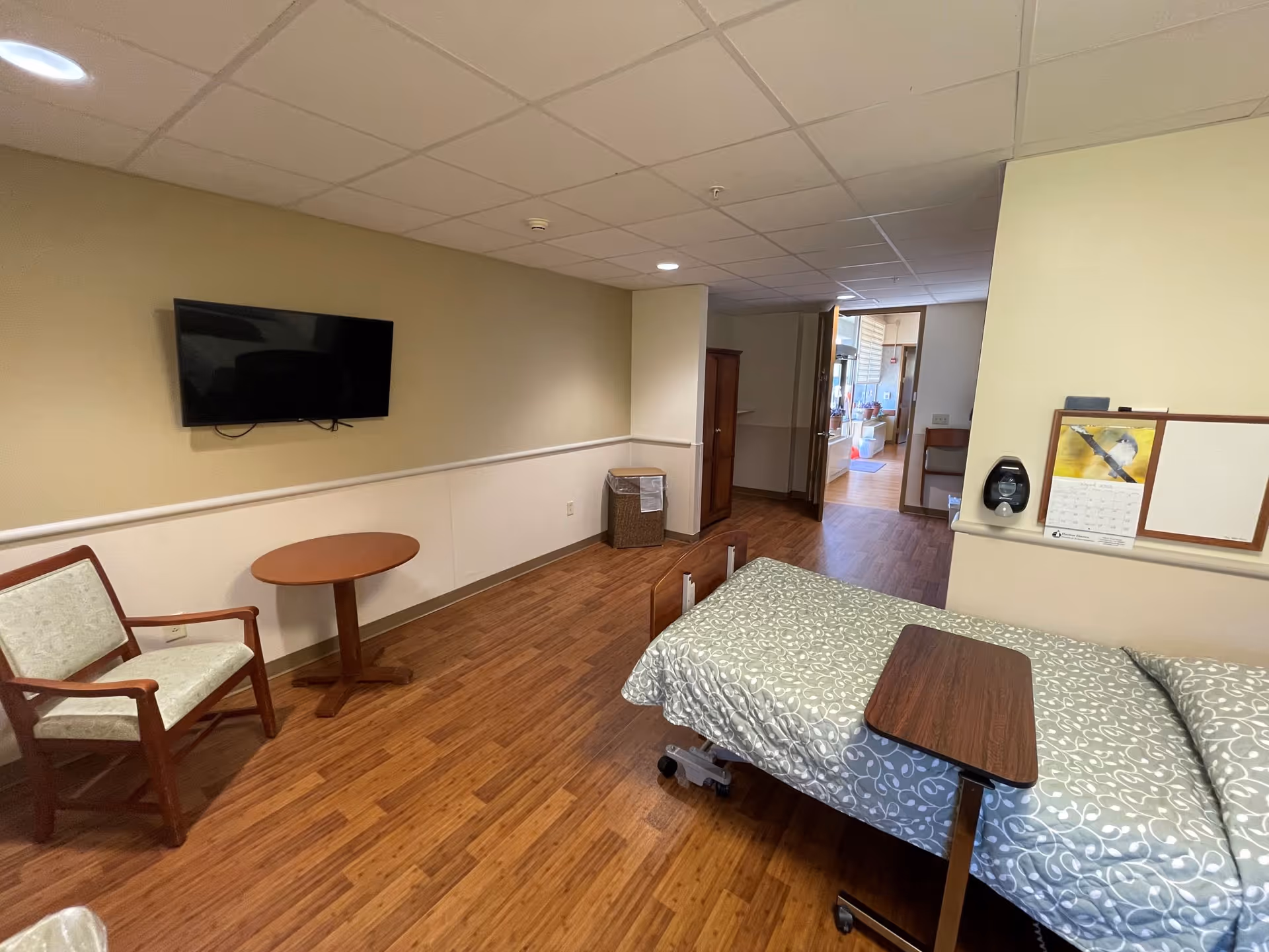 A room in a senior living facility with a single bed covered in a patterned bedspread, a small overbed table, a wooden chair with light upholstery, a round wooden side table, a wall-mounted flat screen TV, a trash bin, and a wooden cabinet. The floor is wood laminate, and the walls are painted beige and white. There is a calendar and a hand sanitizer dispenser on a small ledge near the bed. An open doorway leads to another area of the facility.