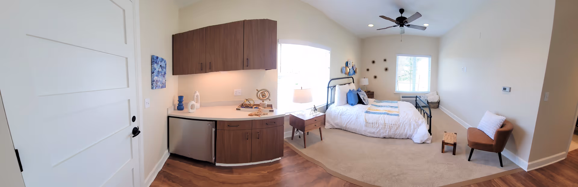 A bright and cozy assisted living bedroom with a white door on the left, wooden cabinets and a small refrigerator below them. The room features a bed with white bedding and blue accent pillows, a wooden nightstand with a lamp, a ceiling fan, a window with blinds, a small brown chair with a white pillow, and a small wooden stool on a beige carpet. The walls are light-colored and decorated with blue and brown wall art.